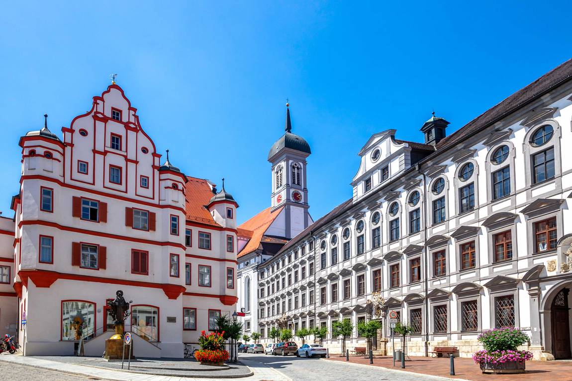 ©Sina Ettmer/stock.adobe.com Eine sonnige Straße in Augsburg, Deutschland, mit dem historischen weiß-roten Perlachturm, verschnörkelten Barockgebäuden, Blumenkübeln und einem strahlend blauen Himmel über der Stadt.