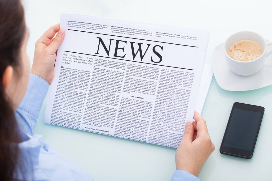 ©Andrey Popov/stock.adobe.com Eine Person hält eine Zeitung mit der Schlagzeile NEWS in der Hand und liest sie an einem Tisch. In der Nähe befinden sich ein Smartphone und eine Tasse Kaffee. Die Person ist von hinten zu sehen.