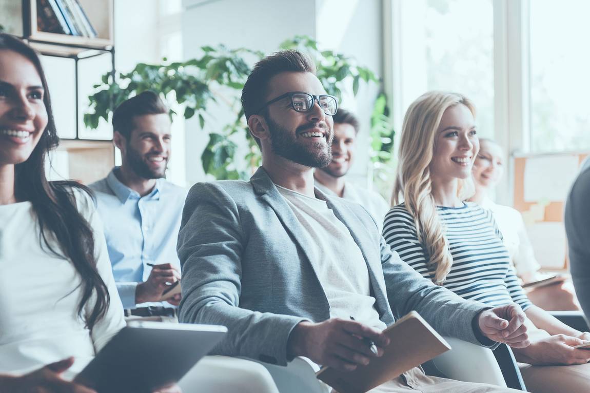 ©istock.com/g-stockstudio Eine Gruppe von Menschen sitzt in einem Innenraum zusammen, lächelt und hört aufmerksam zu. Sie scheinen sich in einer Besprechung oder einem Seminar zu befinden, halten Notebooks und Tablets in der Hand, während hinter ihnen das Sonnenlicht durch große Fenster strömt.